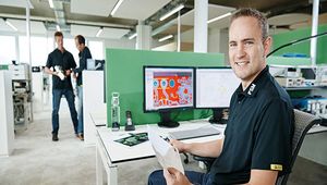 Das Bild zeigt einen Mitarbeiter der FSM AG an einem Arbeitsplatz mit zwei Monitoren, auf denen technische Zeichnungen und Entwürfe zu sehen sind. Im Hintergrund bewegen sich weitere Mitarbeiter durch das Büro. Die Umgebung vermittelt einen Einblick in die moderne Bürogestaltung der FSM AG.