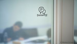 Detailaufnahme einer Glastür mit Piktogramm einer sitzenden Person und Wellenlinien, dahinter ein Besprechungsraum mit einer Person in gestreiftem Oberteil, die verschwommen im Hintergrund zu sehen ist.